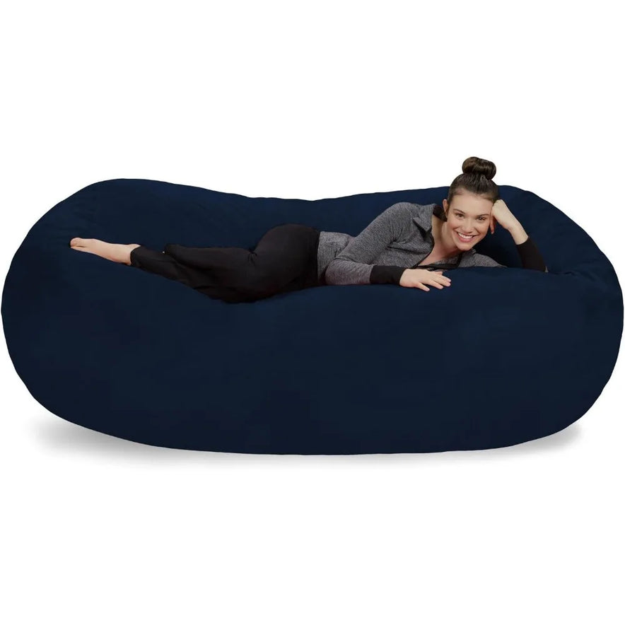 A woman relaxes on a Bean Bag Lounger - Plush Sofas with Soft Microsuede Cover in dark blue, smiling as she rests her head on one hand. She wears black pants and a gray and black jacket.