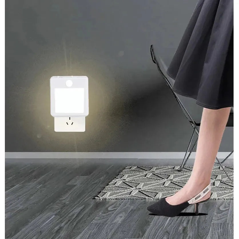 A Dimmable Motion Sensor LED Night Light with EU Plug softly illuminates the wall near a gray rug on the wooden floor. Nearby, a person in a black skirt and low-heeled shoes stands by, with part of a folding chair visible, enhancing an energy-efficient ambiance.