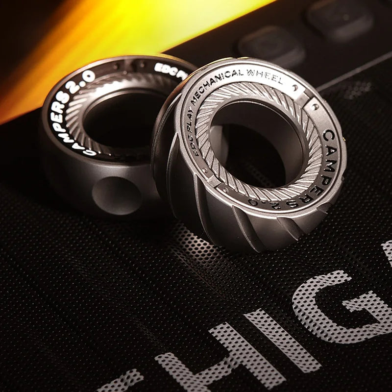 Two metallic, ridged Spystar Mechanic Ring Fidget Spinner Fingertip Gyro Ratchet toys rest on a black textured surface with partial white text, illuminated by warm light—perfect as adult anti-stress office desk EDC.