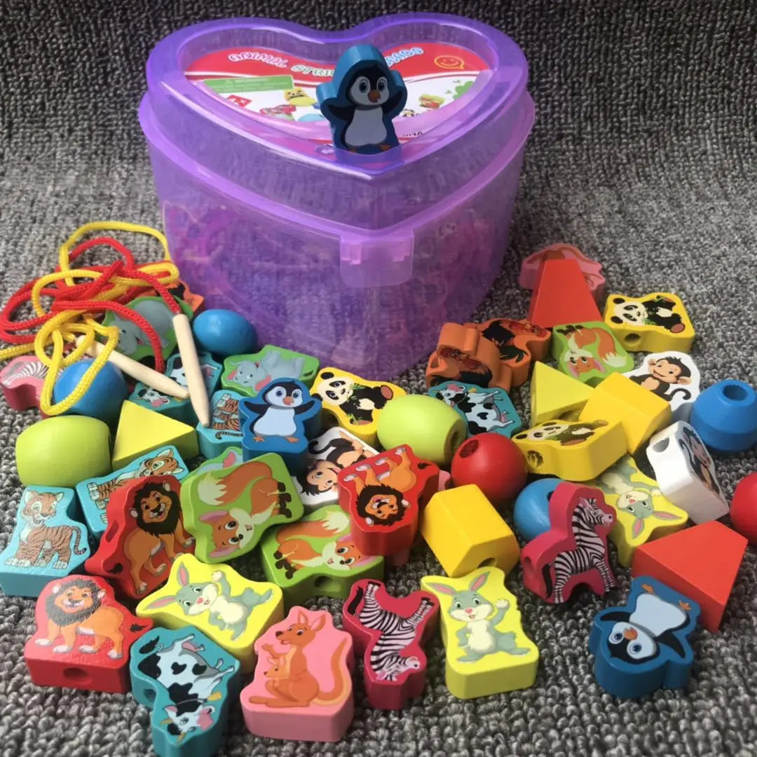 A set of colorful wooden animal and shape blocks lies scattered on a gray surface. A purple heart-shaped container with a penguin figurine is in the background, alongside red and yellow stringing threads with wooden needles, ideal for boosting fine motor skills through educational play using the New 50pcs Baby Wooden Lacing Beads Set.