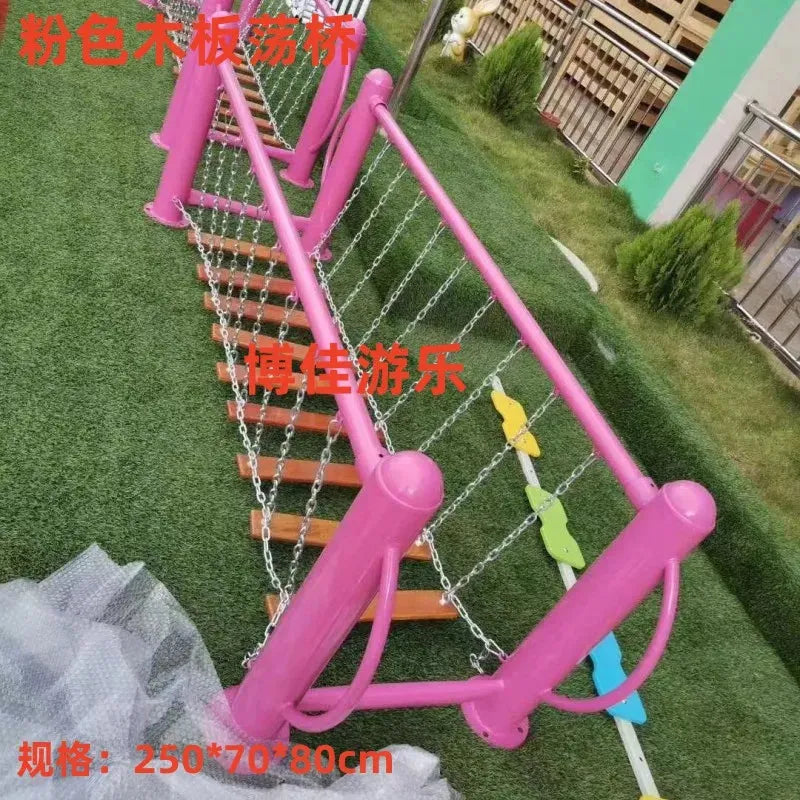 A pink wooden swinging bridge with chains and colorful steps offers adventurous play in this charming playground. As a unisex toy, it spans green grass with fencing behind. Dimensions: 250*70*80cm, making it ideal for young explorers.