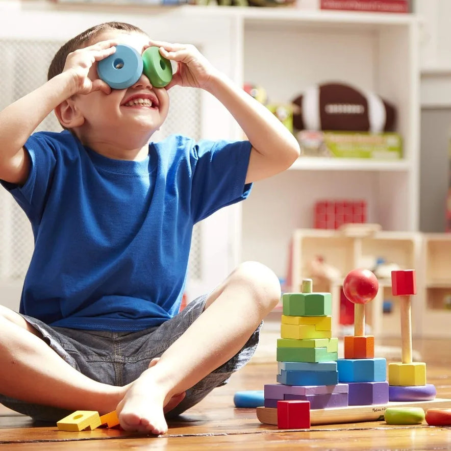 A smiling child in a blue shirt sits on the floor, holding colorful wooden rings from the Melissa and Doug Geometric Stacker Wooden Educational Toy Shape Sorting Cube with 12 Shapes, surrounded by stacked blocks and toys.