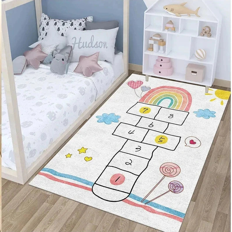 A children’s bedroom features a bed with star and cloud pillows beside a Children’s Play Hopscotch Carpet (Color: 01), an anti-slip large area rug with numbers, rainbows, stars, and lollipops. A white toy shelf stands against the wall.