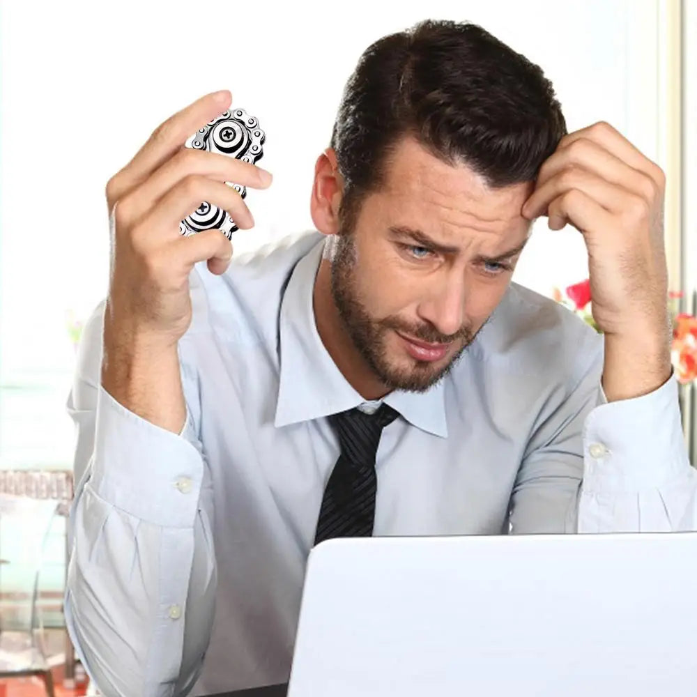A man in a dress shirt and tie looks frustrated at his desk, holding his head with one hand and the Sprockets Flywheel Fidget Spinner in the other, beside an open laptop. The bright room with flowers contrasts sharply with his stressed demeanor as he manages his dropshipping business.