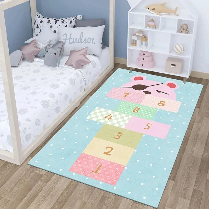 A childrens bedroom featuring the Childrens Play Hopscotch Carpet (Color: 02), a large anti-slip rug with numbered blocks and a bear face. The room also includes a bed, plush toys, pillows, and a white toy shelf against the wall.