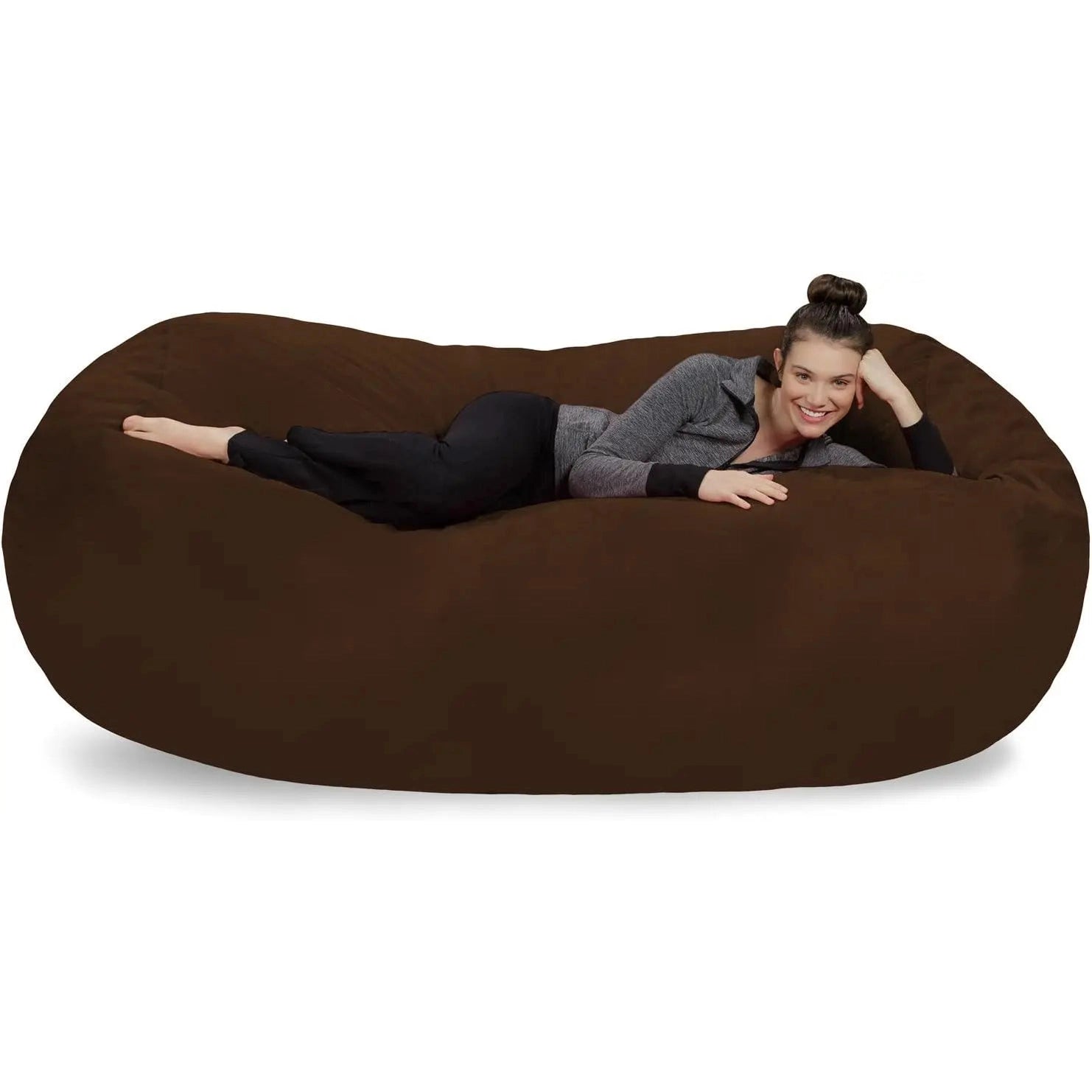 A woman, smiling at the camera, relaxes on a Chocolate Bean Bag Lounger - Plush Sofas with a soft Microsuede Cover. She wears black pants, a gray zip-up top, and has her hair in a neat bun.