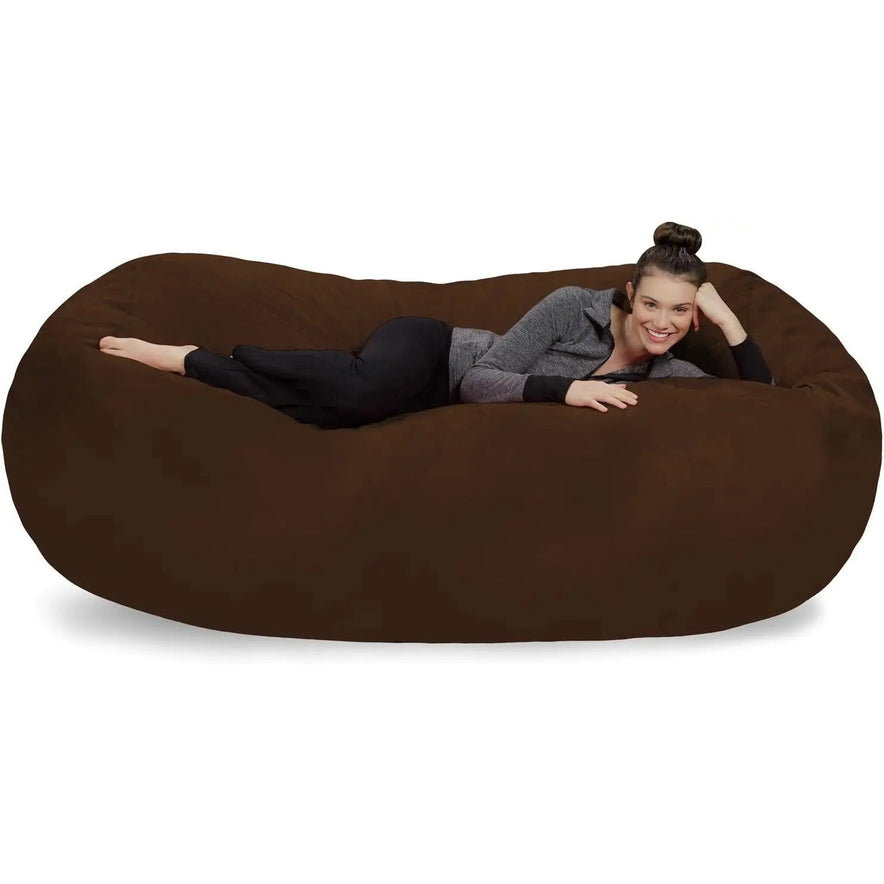 A woman, smiling at the camera, relaxes on a Chocolate Bean Bag Lounger - Plush Sofas with a soft Microsuede Cover. She wears black pants, a gray zip-up top, and has her hair in a neat bun.