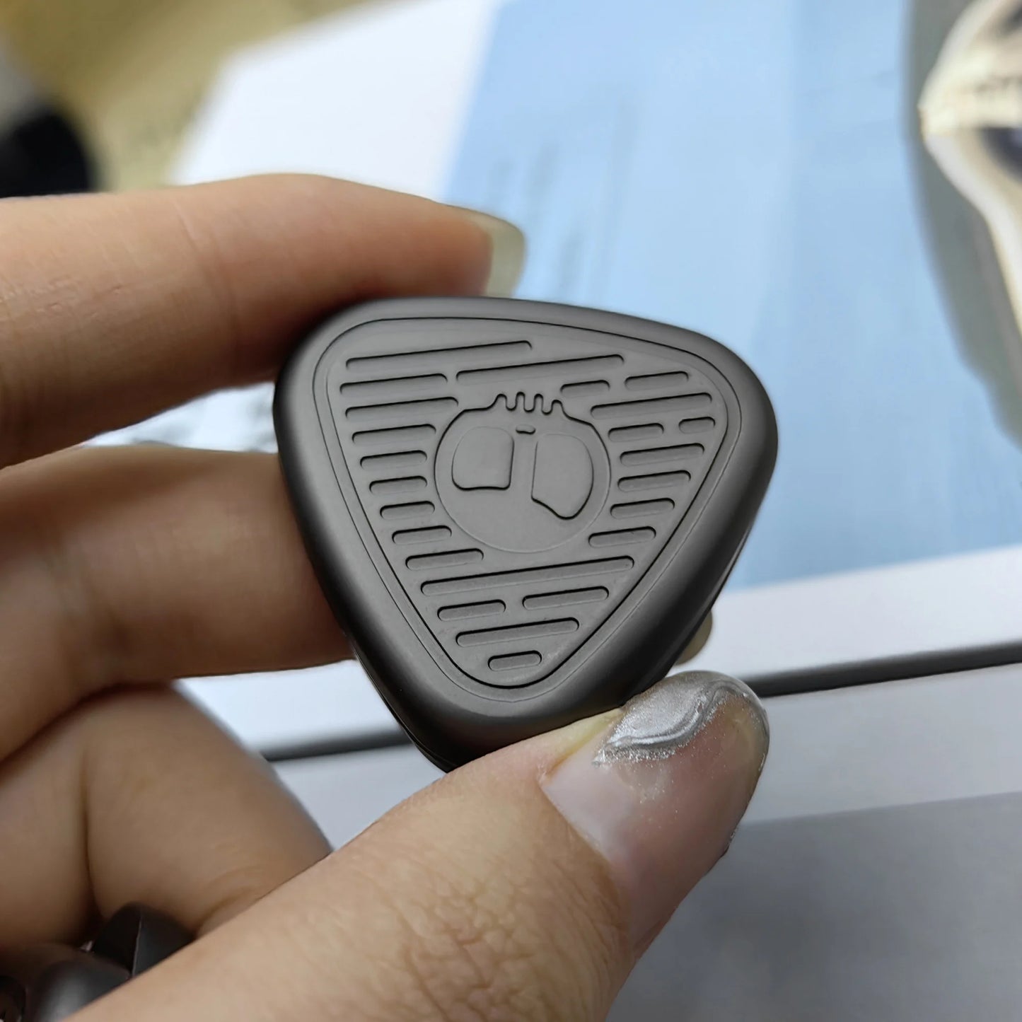 A hand with manicured nails holds a gray EDC Fingertip Toys Fidget Slider—triangular, metallic, with engraved lines and a central circular icon. The slightly blurred background emphasizes the toys sleek finish.