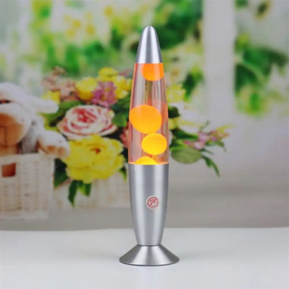 A Mesmerizing Jellyfish Lava LED Night Light, with glowing bubbles and orange liquid, graces a surface. In the blurred background, a floral arrangement with yellow and pink flowers adds a vibrant touch to the scene.