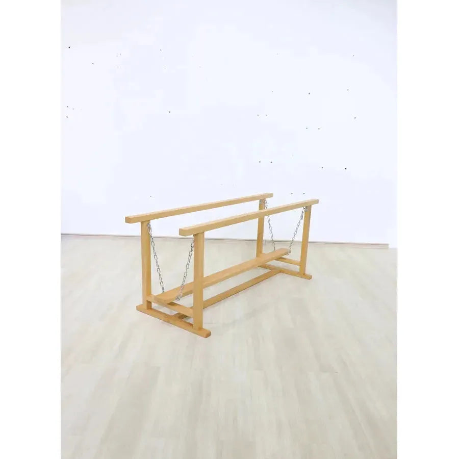 The Indoor Wooden Balance Beam Sensory Training Toy for Children 3-12 Years is a stylish wooden bench-like structure with a smaller bench below, suspended by chains. Set on a light wood floor against a white wall, it offers sensory system training potential.