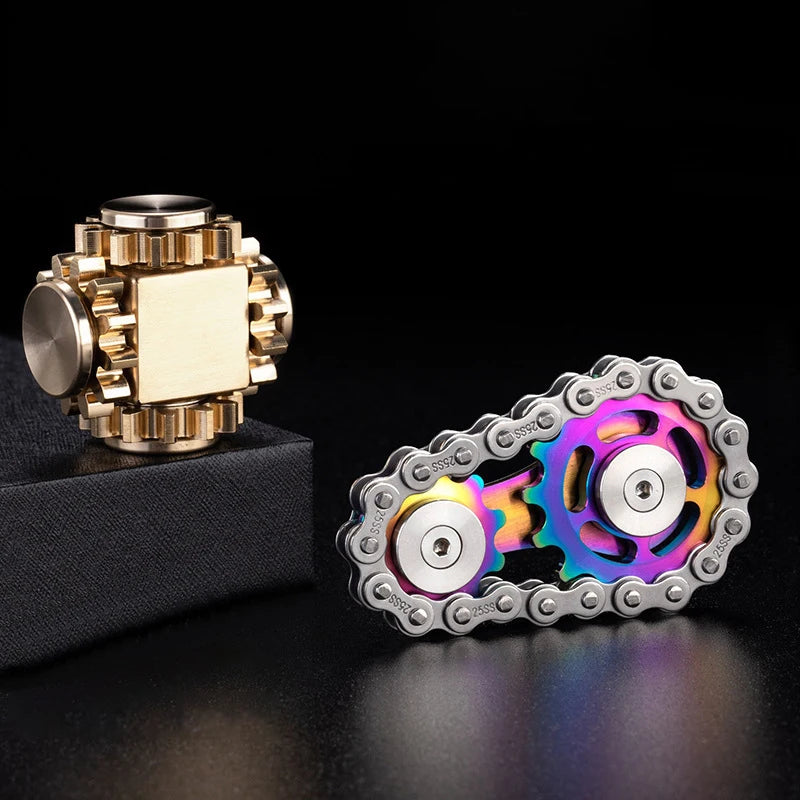 Two metal fidget toys rest on a dark surface: one resembles intertwined gears with a metallic finish, while the other is a multi-colored Bike Chain Metal Fidget Spinner with Magnetic Slider, featuring circular components for stress relief and fun.