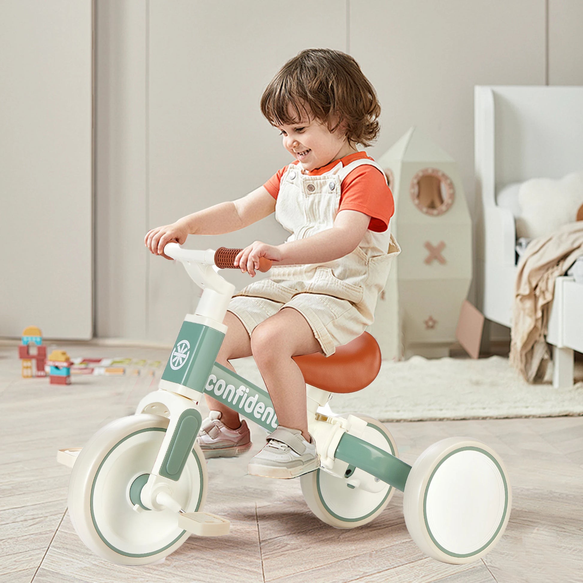 A young child smiles while riding the Baby Balance Bike for Kids 1 to 3 Years Old Toddler Trike with Parent Steering Push Handle, wearing beige overalls and an orange shirt in a playroom filled with toys and neutral-colored furniture.