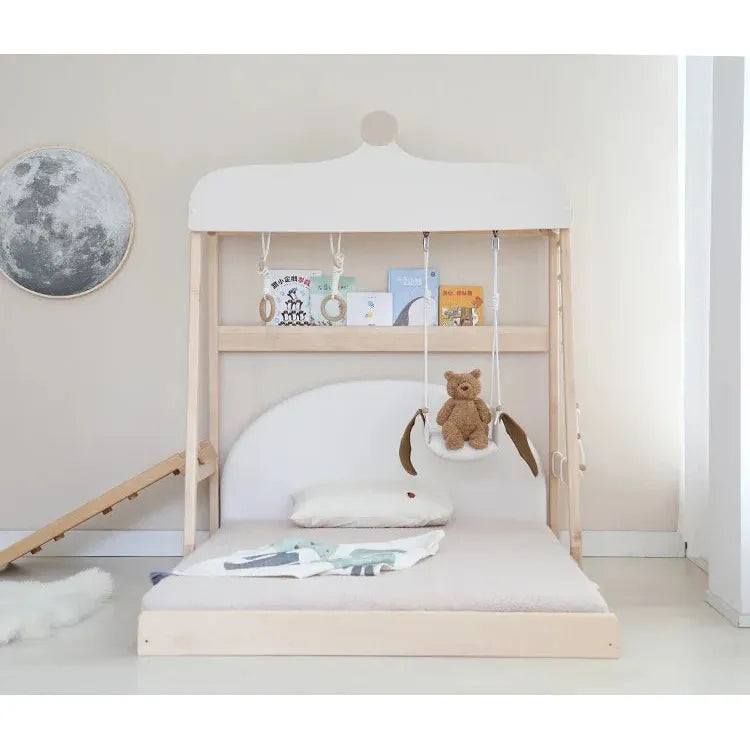 The Versatile Solid Wood Childrens Bed boasts a white canopy and small slide. Above the bed, books sit on a shelf while a teddy bear swings from ropes. A round moon decor adorns the beige wall, adding charm to this cozy space.