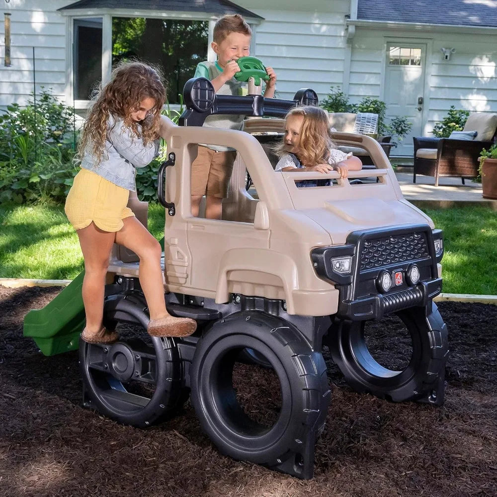 Three children play on the Truck Climber Kids Outdoor Playset with Slide and Wall: one climbs the wall, another steers from the top, and a third sits inside. A house is visible in the backyard background.
