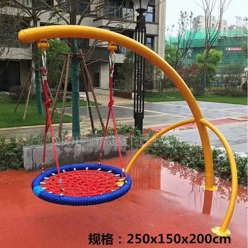 A childrens playground features an Outdoor Steel Climbing Frame Swing for Sensory Integration Training, with a circular rope swing sporting a blue rim and red netting, hung from a yellow arch frame. The area has red flooring amidst grass and trees, with buildings in the background.
