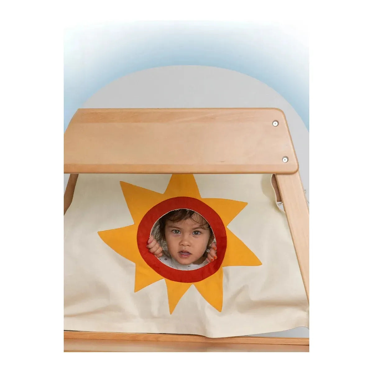 A child peers through a sun-shaped circular cutout in the fabric panel of the Indoor Adventure Climbing Frame for Kids with Slide and Swing, resembling a sunny day playhouse. Its attached to a wooden frame, evoking an indoor playground tent, set against a light blue gradient background.