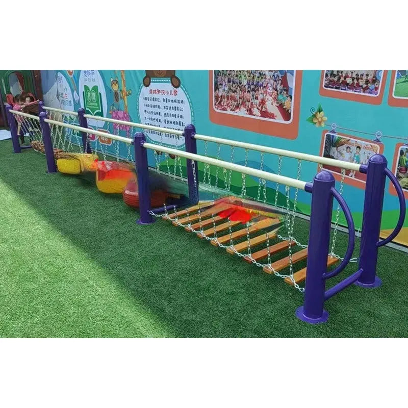The childrens outdoor playground includes a climbing net and swinging bridge made of wooden planks and chains for unisex play, colorful rope buckets fun for ages 3-8, all set on artificial grass surrounded by a vibrant mural wall with playful metal toys.
