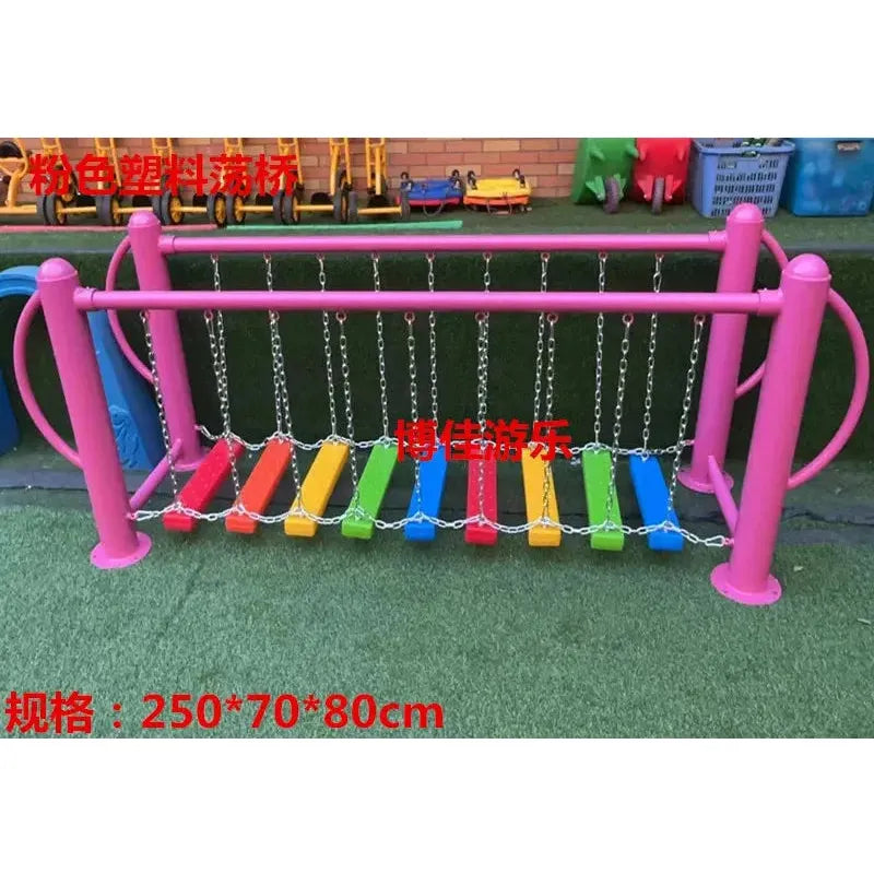 A playground bridge features pink plastic boards and is set on a green surface with scattered toys. Metal toys and equipment are in the background, and red measurements are visible on the image.