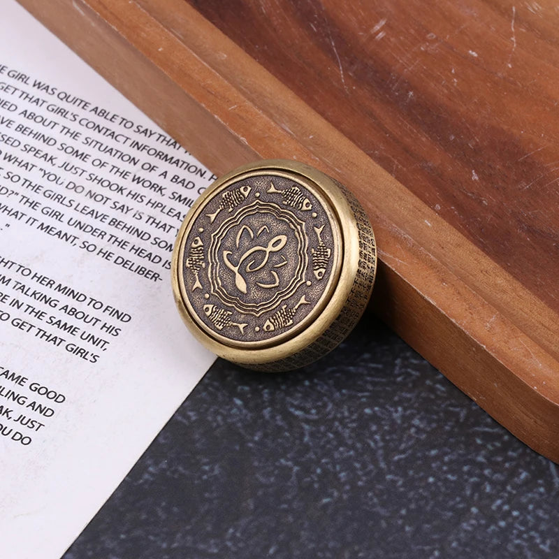 A round, antique-style brass compass with detailed engravings rests on a book and wood, evoking the calming effect of the Zen Buddha Fidget Spinner Metal EDC Fidget Toy for Adults Stress Relief Anxiety Hand Spinner. The lid has decorative symbols and patterns.