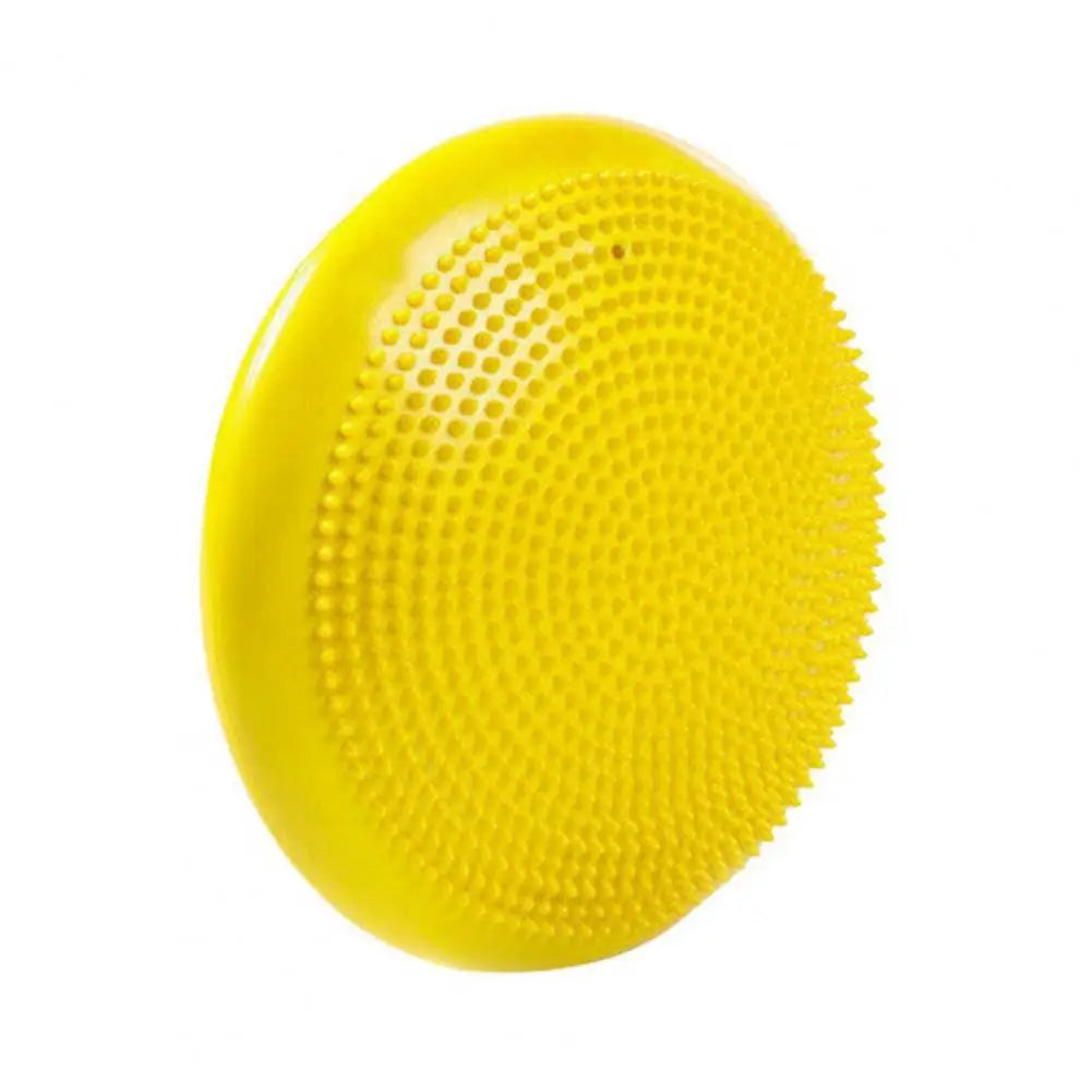 A yellow circular silicone scrubber, resembling a balance disc, features a textured surface with small bristles for gentle cleansing. Set on a flat base, it is captured against a plain white background.