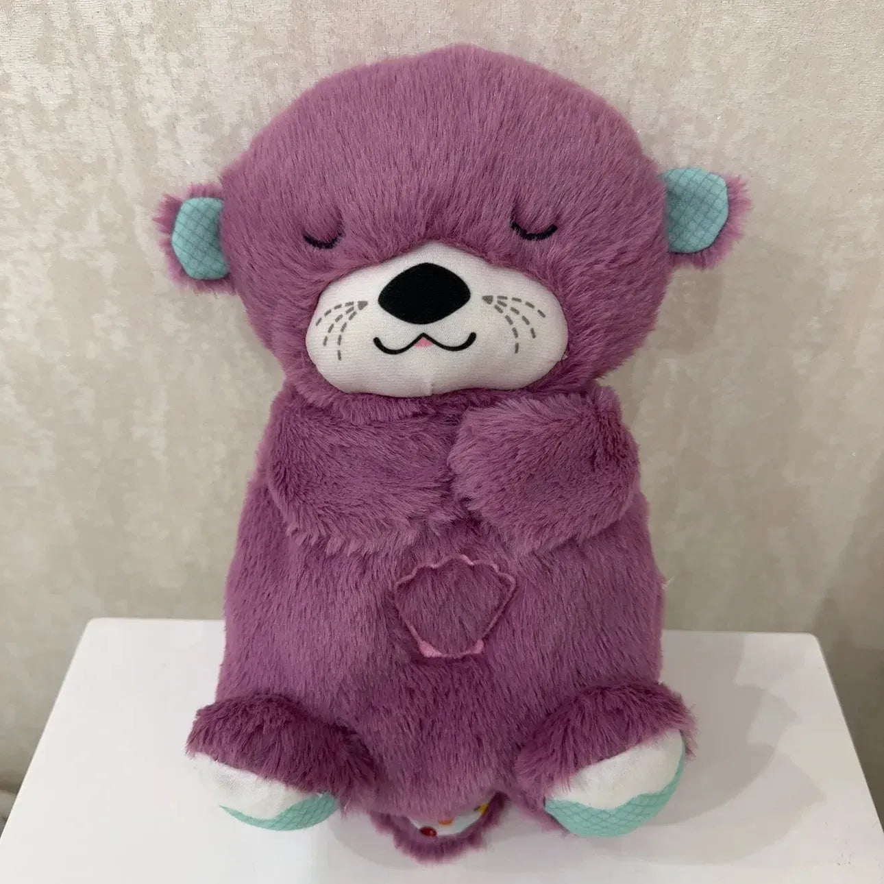A purple Soothing Otter Musical Plush Toy for newborns sleeps on its back with closed eyes. Its white face and paws, light blue inner ears, and a small heart on its chest create a cozy scene against a neutral wall, making it an ideal sensory sleep companion and comfort gift.