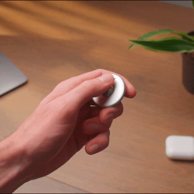 A hand spins the ONO Scroller Aluminum fidget keychain on a wooden desk, with a plant, part of a laptop, and white wireless earbuds visible in the background.