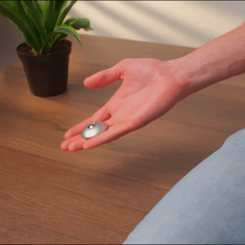 A hand gently tosses the ONO Scroller Aluminum—a sleek, stone-like steel rollerball—above a wooden table, with a green potted plant in the background.