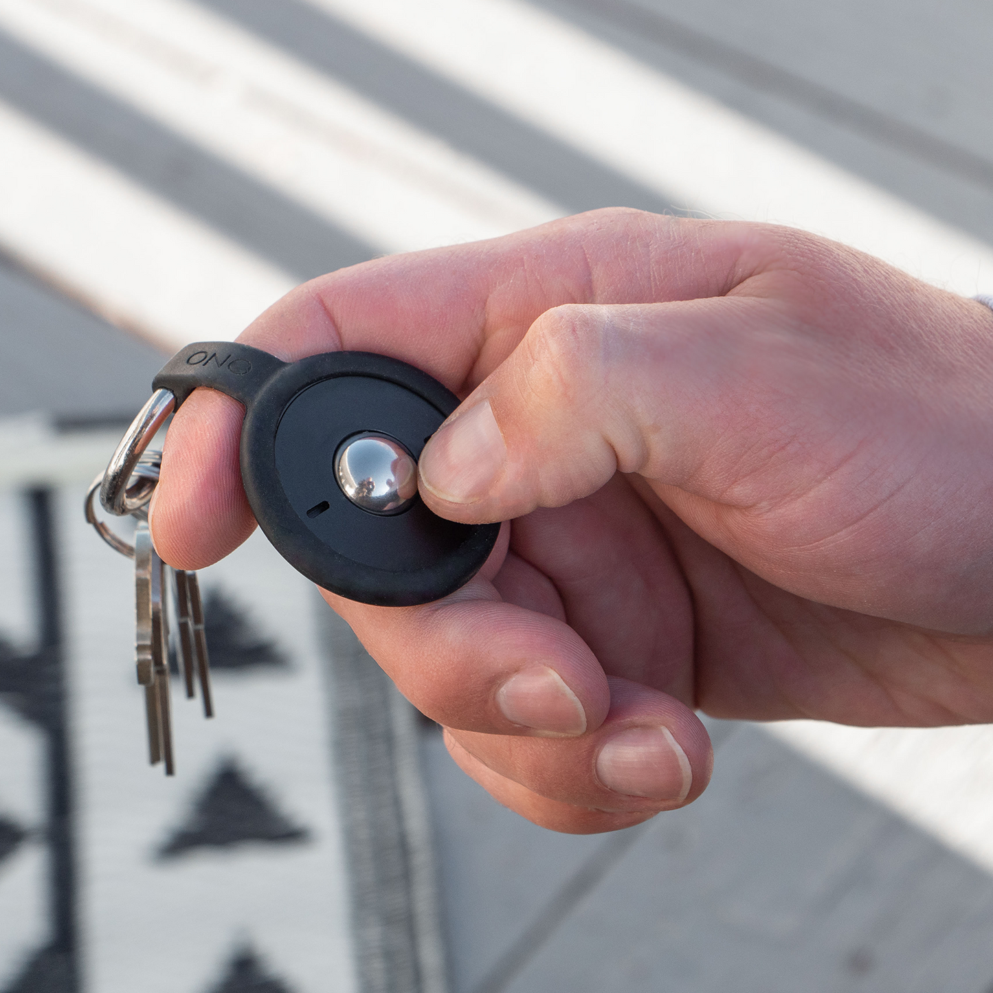 A hand holds a set of keys with the ONO Scroller Keychain, a round black device with silicone keychain, thumb pressing the button over a patterned rug background.