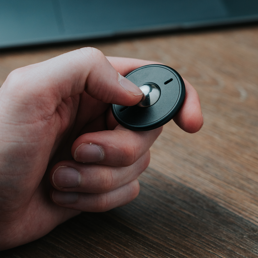 A hand holds an ONO Scroller Aluminum, a small, round fidget keychain with a silver spinning center, on a wooden surface with part of a laptop in the background.