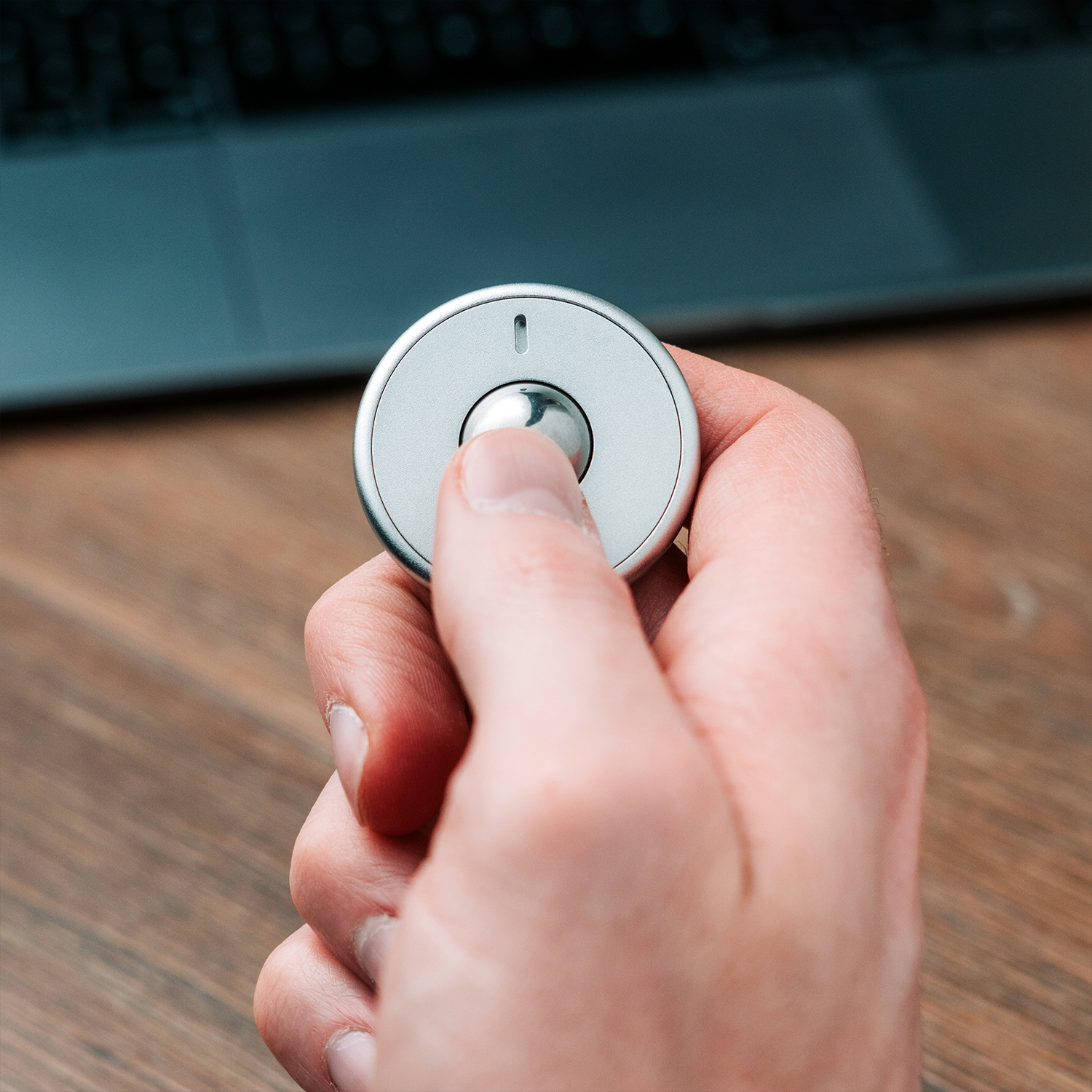 A hand presses the small, round ONO Scroller Aluminum wireless remote control, featuring an aluminum scroll wheel, with a blurred laptop keyboard in the background on a wooden surface.