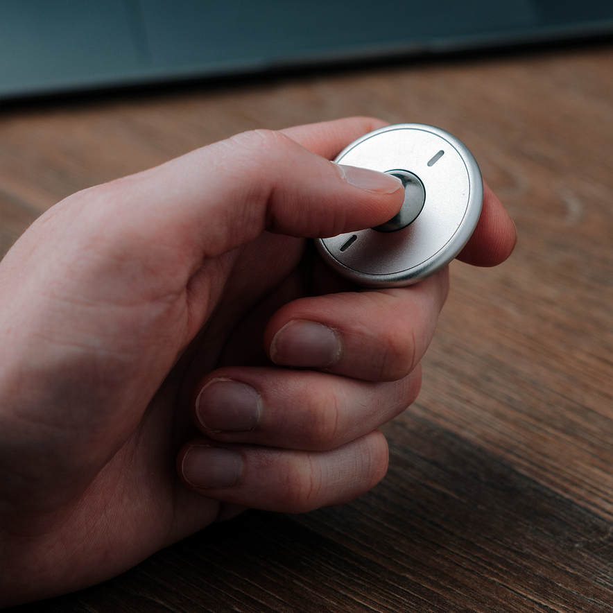 A hand holds the ONO Scroller Aluminum, a small round metallic fidget spinner with an aluminum scroller button, above a wooden surface with a laptop partially visible in the background.