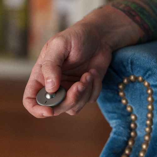 A close-up of a hand holding the ONO Scroller Titanium fidget toy with a button, resting on the edge of blue upholstered furniture accented with decorative brass studs.