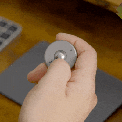 A hand rotates the ONO Scroller Titanium—a gray, circular wireless controller with a metal knob—over a desk with a tablet and keyboard, making it perfect as both a computer input device and a fidget toy.