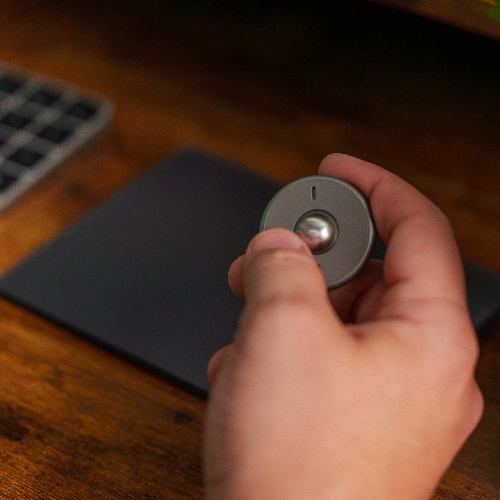 A hand holds the ONO Scroller Titanium, a sleek gray circular device with a central knob, over a dark touchpad on a wooden desk, with part of a keyboard visible in the background.