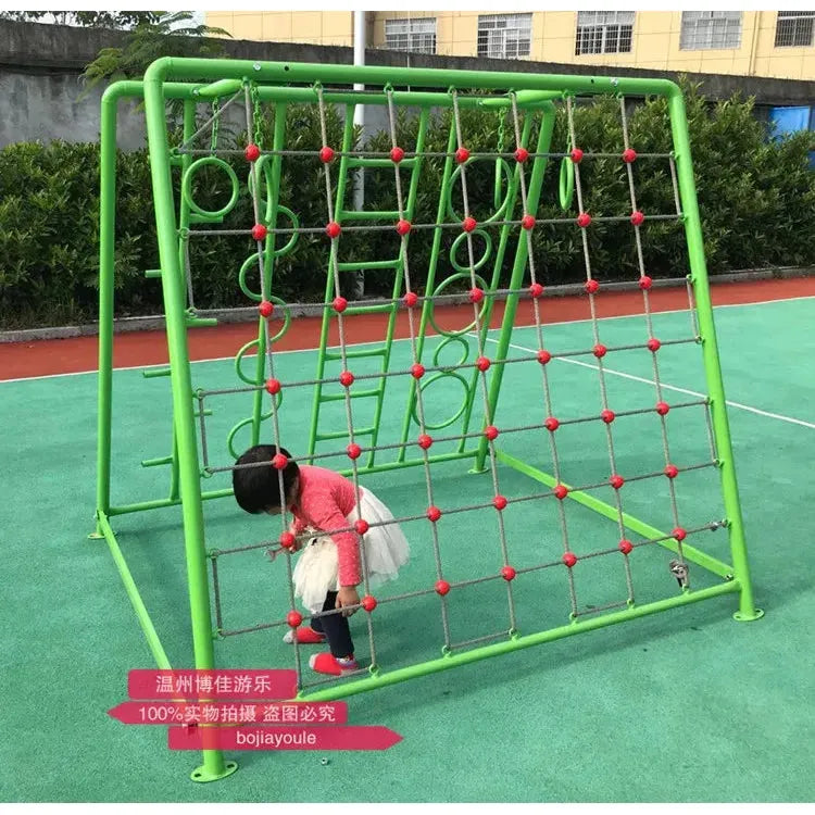 A child in a red top and white tutu joyfully plays on an outdoor toys climbing net with red ropes and various rings, suitable for ages 3-8. This unisex sports equipment sits on a paved area with green bushes as a lush backdrop.