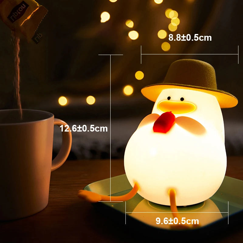 A Cute Cartoon Duck Rechargeable LED Night Light, perfect for kids room decor, emits a cozy glow. The silicone lamp sits on a green plate, measuring 8.8x9.6 cm. Nearby, coffee or tea fills a mug of 12.6 cm as warm yellow lights enhance the ambiance with the night lights gentle illumination.