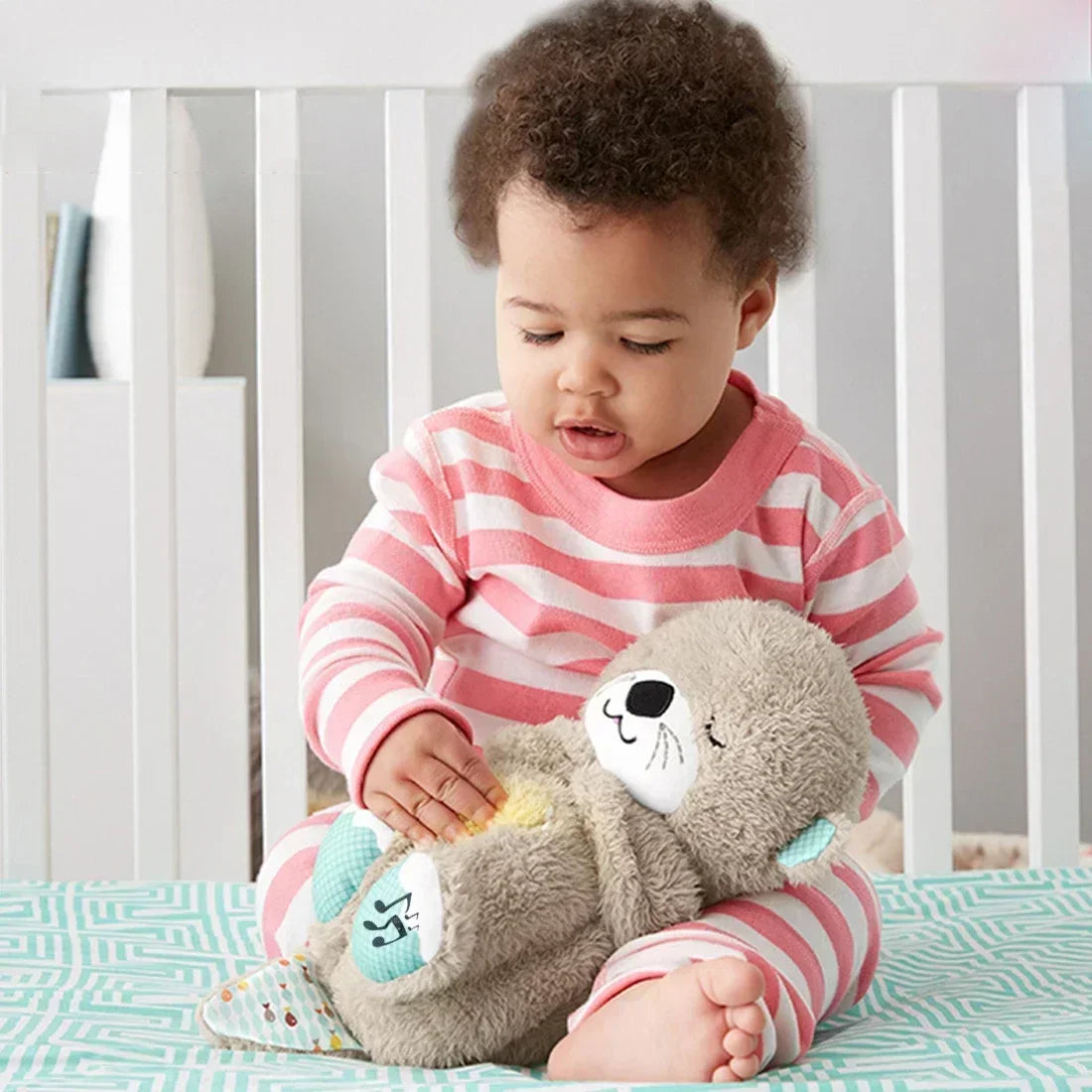 A baby in pink and white striped pajamas sits on a patterned blanket, playing focused and content with the Soothing Breathing Otter Musical Plush Toy. The toy offers rhythmic comfort amid a white crib set quietly in the background.