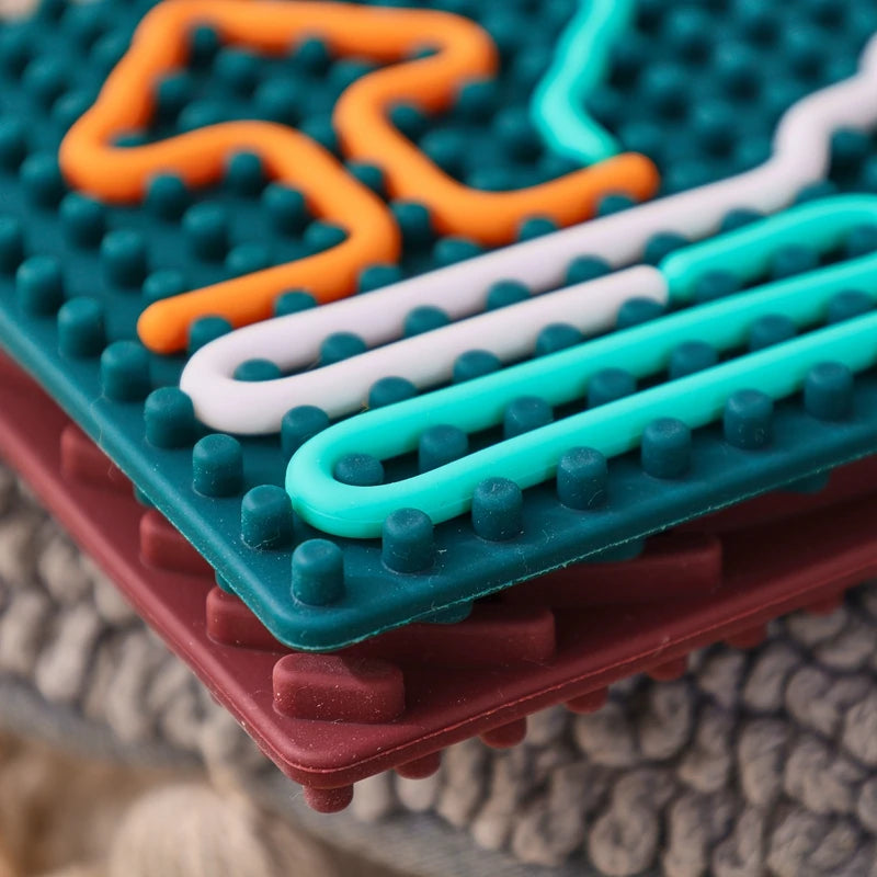 Close-up of two stacked Silicone Montessori Sensory Activity Board Fidget Toys for Kids Anxiety Relief and Mindfulness, featuring raised bumps and colorful wavy lines. One mat is teal, the other maroon, on a textured gray fabric surface.