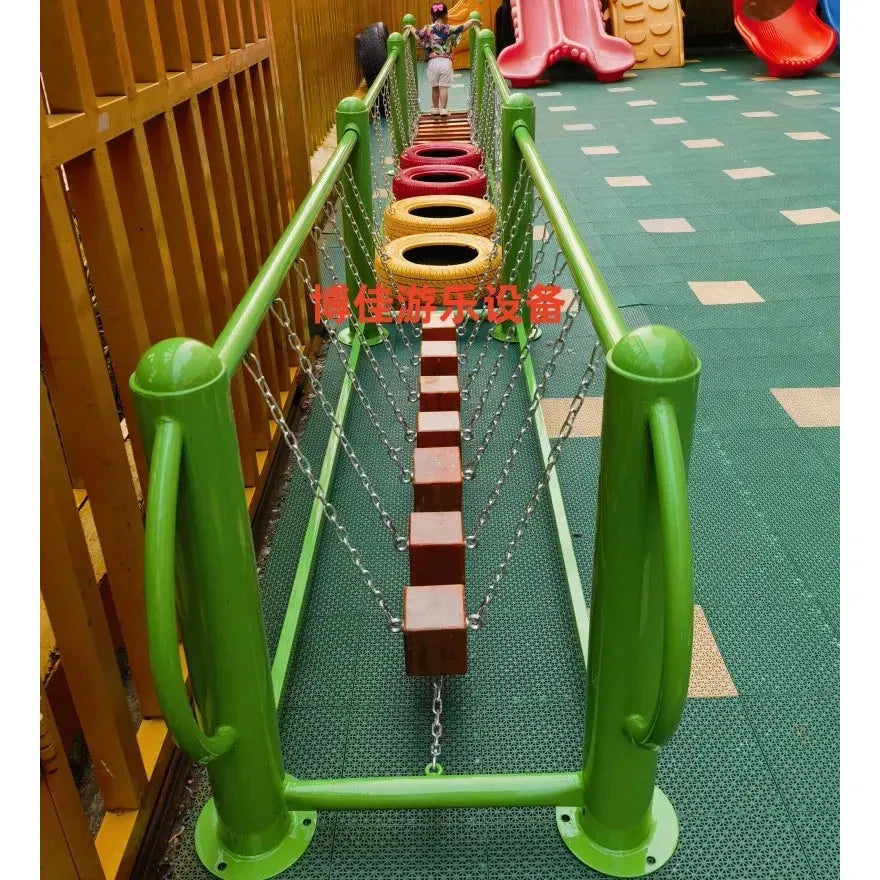 A fenced area features a green swinging bridge with chain-linked wooden planks and circular elements on a rubber mat. Nearby, colorful slides and childrens toys from the Outdoor Toys collection enhance the playful atmosphere designed for kids activities.