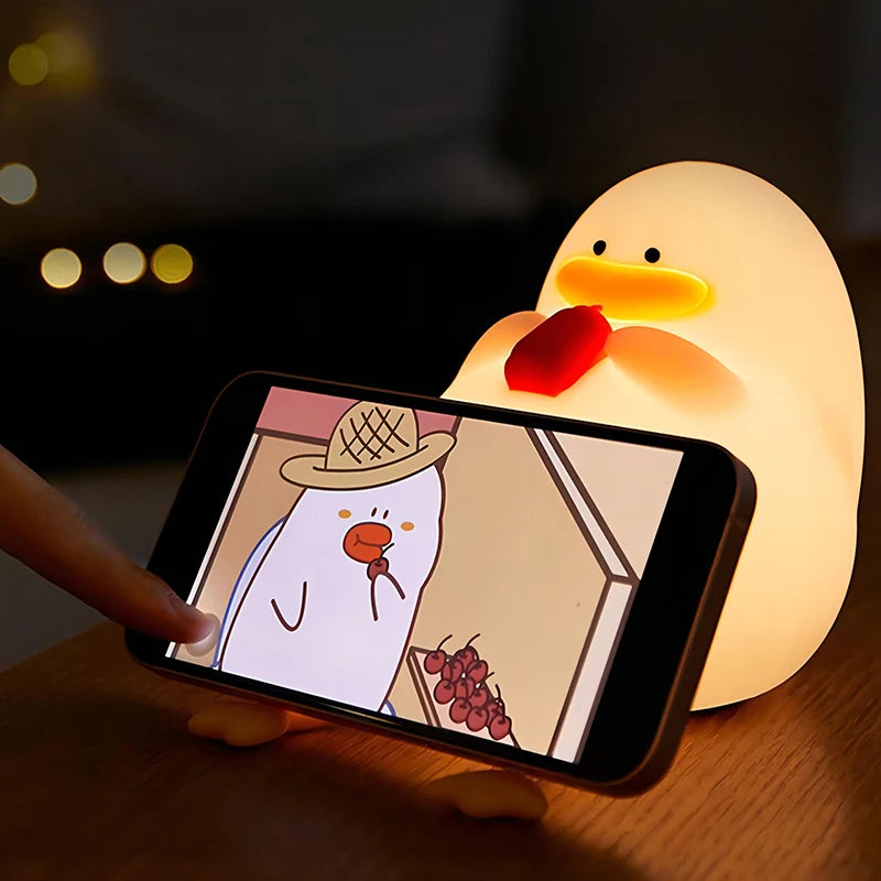 The Cute Cartoon Duck Rechargeable LED Night Light, a silicone lamp for kids room decor, gently glows in a dimly lit room. A smartphone leans against the portable light, displaying a cartoon duck with a detective hat holding grapes, as a finger touches the screen.