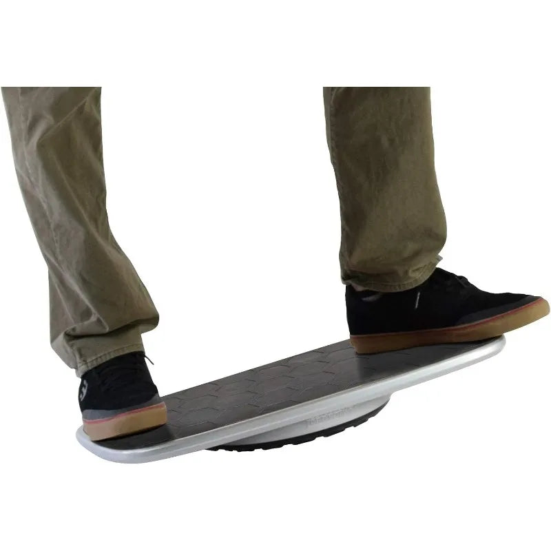 A person wearing khaki pants and black sneakers balances on the BASE Desk Standing Board, a black and silver wobble board with an anti-fatigue mat and rounded base, to promote better posture while standing.