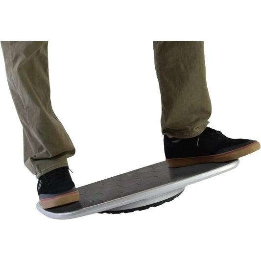 A person wearing khaki pants and black sneakers balances on the BASE Desk Standing Board, a black and silver wobble board with an anti-fatigue mat and rounded base, to promote better posture while standing.