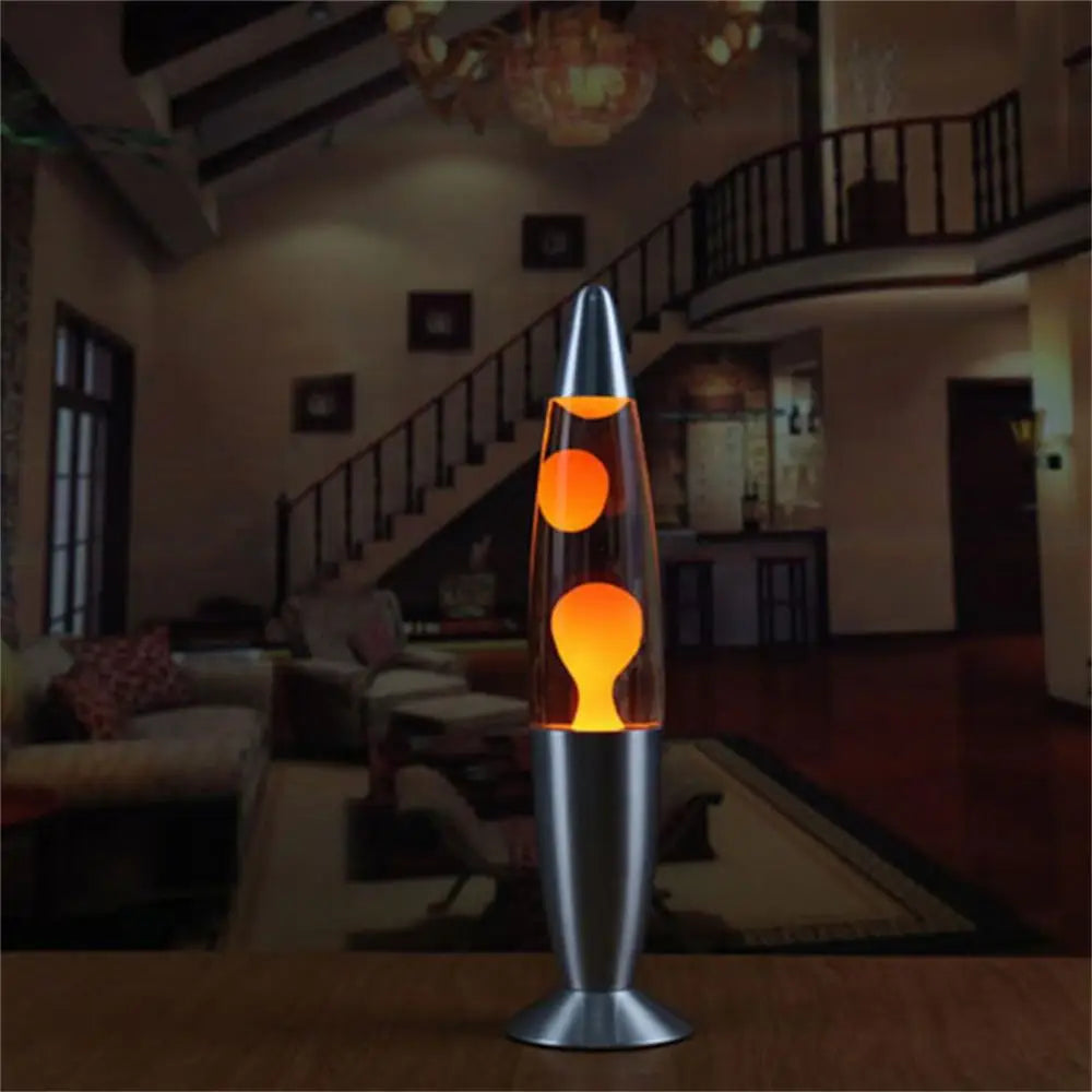 A Mesmerizing Jellyfish Lava LED Night Light with orange liquid and floating blobs sits on a wooden surface. Behind it, a dimly lit living room displays a staircase, chandelier, and sofas, offering the ideal cozy ambiance for this captivating aluminum alloy lamp in kids rooms or home settings.