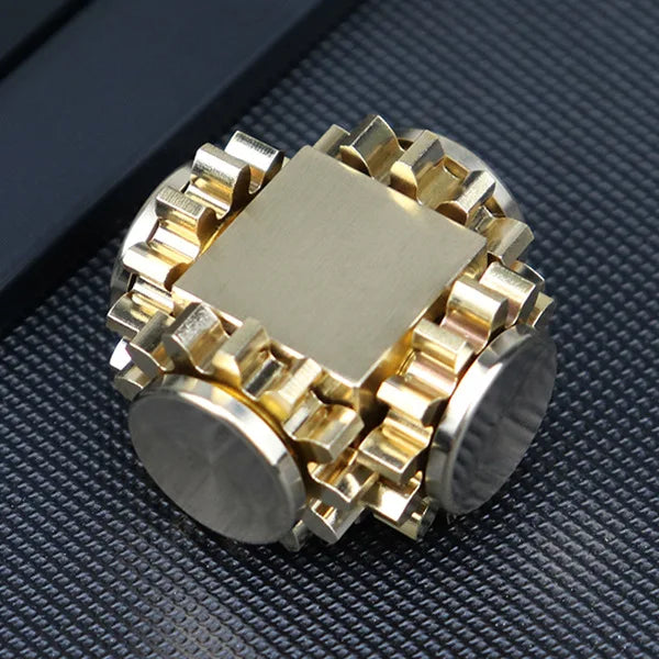 A gold and silver gear-like fidget cube sits on a textured dark surface, its intricate interlocking gears resembling the Bike Chain Metal Fidget Spinner with Magnetic Slider, featuring smooth ends and a mechanical design for stress relief with every twist.