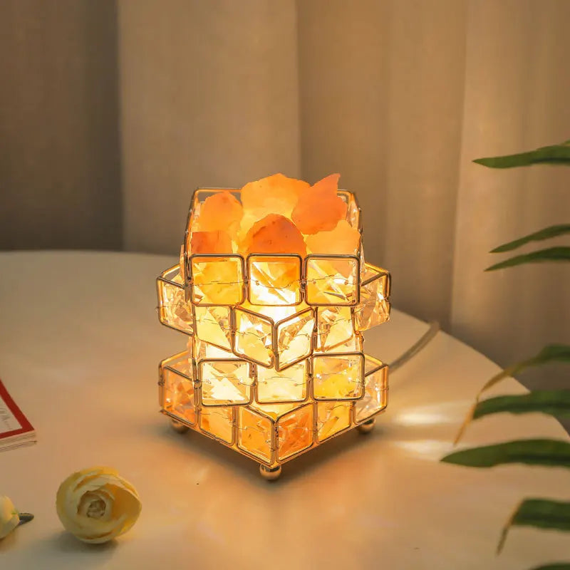 The Himalayan Salt Crystal LED Lamp emits a warm glow, creating a cozy ambiance with its square rocks in a geometric metal frame. A yellow rose and plant enhance the charm, and its USB plug makes it convenient for any setting.