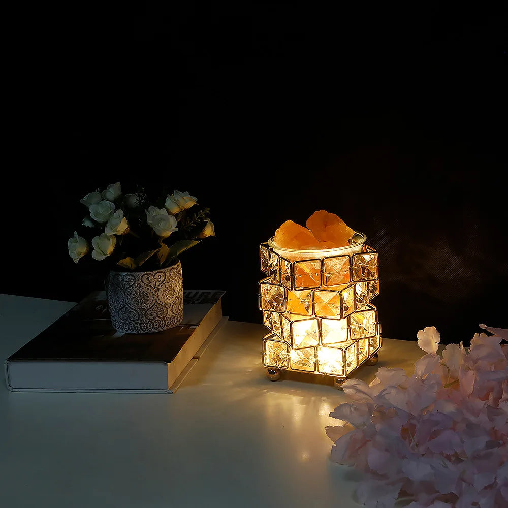 The Himalayan Salt Crystal LED Lamp - USB Night Light for Home Décor and Relaxation, featuring a geometric design, sits beside a flower pot and books. Pink petals adorn the foreground, while the dark background enhances the lamps warm glow, providing easy lighting with USB plug convenience.
