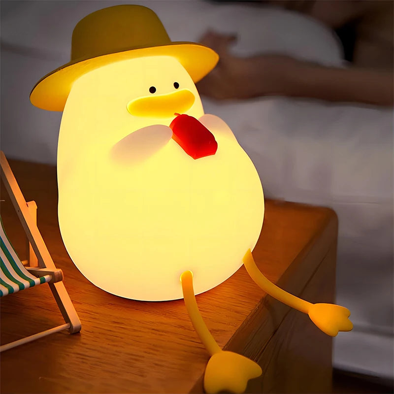 The Cute Cartoon Duck Rechargeable LED Night Light, featuring a silicone duck with a yellow hat and dangling legs, sits on wood emitting a warm glow. A persons arm is visible on a bed in the background.