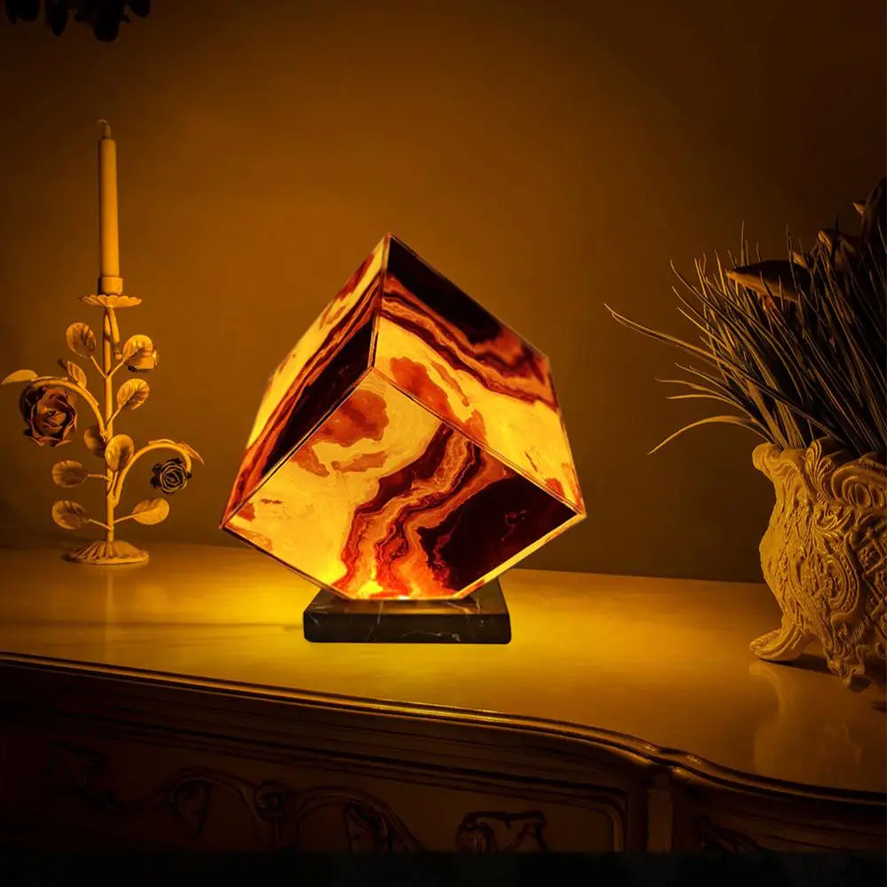 The Elegant Agate Crystal Table Lamp with a marbled brown and beige design sits on the table, flanked by an artistic flower-shaped candleholder and a plant-filled decorative vase. Its warm amber glow perfects the cozy bedside decor ambiance.