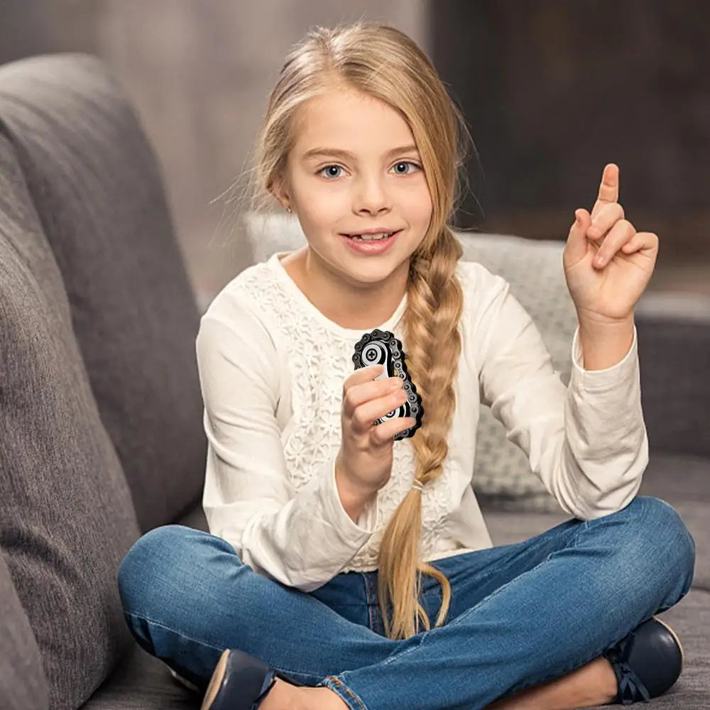 A young girl with long blond hair sits cross-legged on a sofa, smiling while holding a Sprockets Flywheel Fidget Spinner. Shes wearing a white long-sleeve shirt and blue jeans, pointing upwards as if inspired by MezoJaoies latest dropshipping trend.