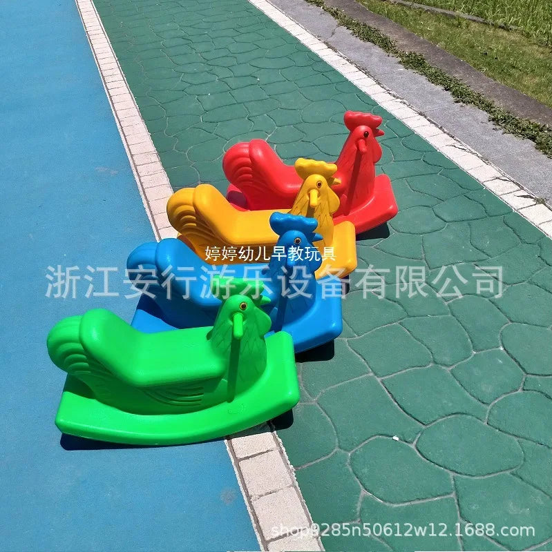 Four vibrant plastic rooster-shaped rocking toys for toddlers, ideal as gifts, are arranged on a playground. They come in green, yellow, red, and orange and sit on a blue and green rubber safety surface.